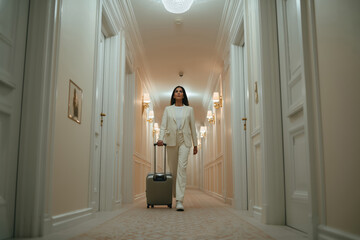 Young adult Caucasian woman walking down hotel hallway pulling suitcase, wearing business attire, looking forward with confident expression, representing hotel service and hospitality