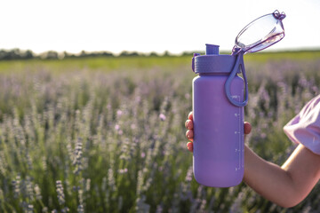 Child’s hand holding a purple water bottle in a blooming lavender field on a sunny day. Hydration and summer lifestyle concept with pastel tones and nature background
