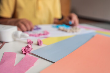 Close-up of a child's art table with colorful pastel paper, pink paper scraps, glue, and a handmade...