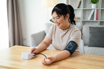 An Asian woman uses a portable blood pressure monitor in her living room. Health awareness concept.