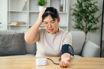 An Asian woman uses a portable blood pressure monitor in her living room. Health awareness concept.
