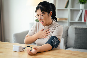An Asian woman uses a portable blood pressure monitor in her living room. Health awareness concept.