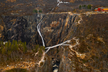 Voringsfossen waterfall cascades down rugged dark rock face with modern bridge crossing over canyon. Dramatic scale of natural wonder in Norway
