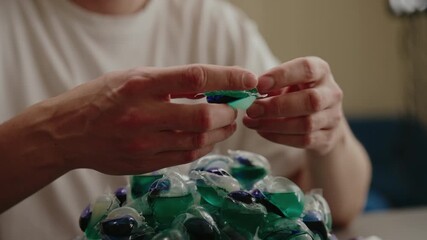 Close-up of man carefully examining laundry detergent pod with pile of similar pods blurred in foreground sitting at table preparing to wash clothes. Concept of convenient washing solutions. - Powered by Adobe