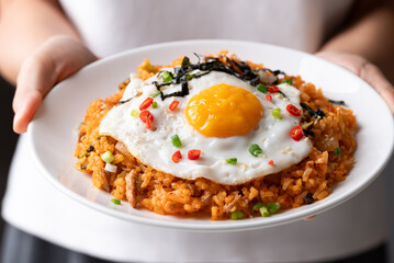 Korean kimchi fried rice with fried egg, pork, sesame seed, chili, spring onion and seaweed on plate holding by hand