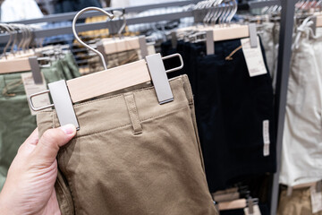 Hand Holding Khaki Pants in Clothing Store Display. The background shows various neatly displayed pants in multiple colors, emphasizing retail fashion, shopping experience, and consumer lifestyle.