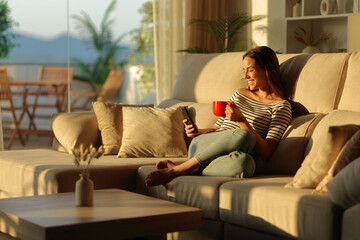 Happy woman using smart phone drinking coffee from red cup at home