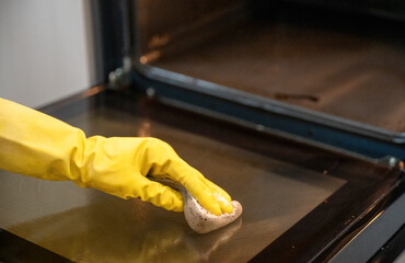 Person cleaning oven door glass with sponge with yellow rubber gloves