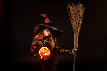 Young woman wearing halloween costume witch on black background holds pumpkin jack o lantern and...