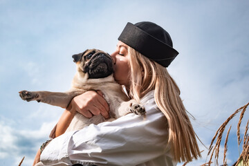 Young happy beautiful woman holding, kissing and hugging her beloved dog during a walk with harness...