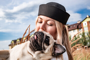 Young happy beautiful woman holding, kissing and hugging her beloved dog during a walk with harness and black leash in a city park. Street photography.	