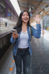 Young woman waving goodbye at train station with smartphone