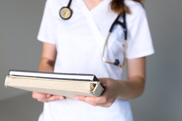 A healthcare worker stands in a clinic, holding two textbooks closely. The individual wears scrubs...