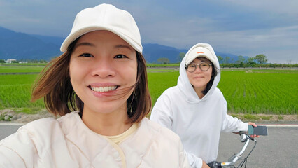 A young lesbian couple smiling cycling through Mr. Brown Avenue in Chishang, Taitung, Taiwan —...