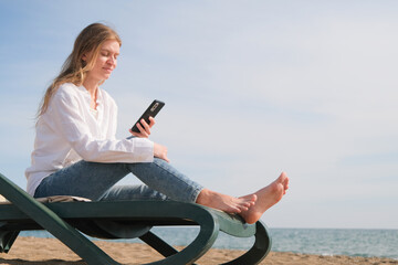 young woman sitting in the chaise lounge on the edge of the sea using smart phone