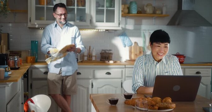 A successful Asian businesswoman works on a laptop during breakfast while her Caucasian husband helps her in the kitchen. Multiracial couple shares morning routines before work at home in the kitchen.