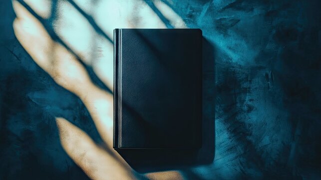 A dark-covered hardcover book rests on a textured, teal surface, bathed in dramatic, contrasting light and shadow patterns cast from an unseen source - Powered by Adobe