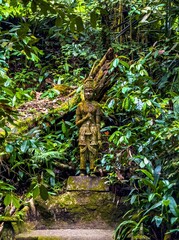 A weathered, moss-covered stone statue of a traditional Thai deity stands majestically on a mossy stone pedestal, nestled deep within a vibrant, dense tropical jungle. The statue, captured in a medium