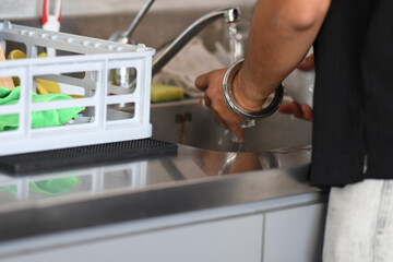 A person's hands are seen washing a glass under running tap water in a modern stainless steel kitchen sink with a dish rack to the left