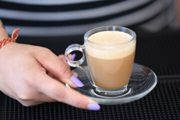 Close up of a barista's hand holding a clear glass cup filled with frothy creamy coffee with a delicate foam topping served on a saucer