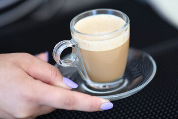 A Person's Hand Serving a Delicious Hot Coffee in a Clear Glass Cup with Frothy Cream on Top and a Saucer on a Dark Background with Soft Lighting