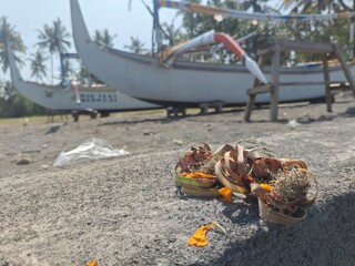 A close-up photo focused on canang sari, a traditional Balinese offering, placed on a rock near fishing boats under sunlight, symbolizing ritual life.

