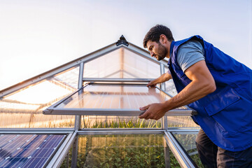 Solar greenhouse installation. A man installs a solar panel on a greenhouse roof. Renewable energy, sustainable agriculture, and eco-friendly technology concept.