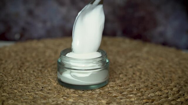 Transferring homemade natural face or body cream into a transparent glass jar. Clean beauty and eco-friendly skincare concept on beige and dark background with a minimalist style.