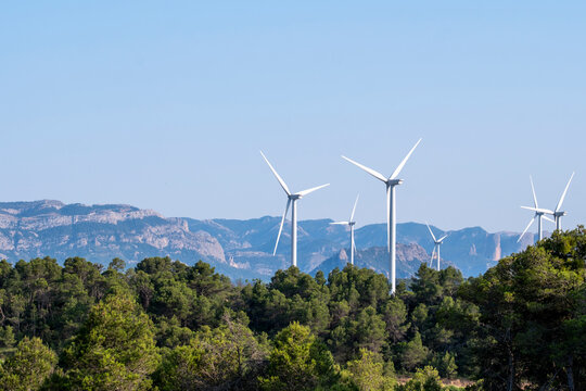 Row of modern wind turbines surrounded by green forest hills under blue sky, Terra Alta, Catalonia, Spain, symbolizing sustainability and wind power production.