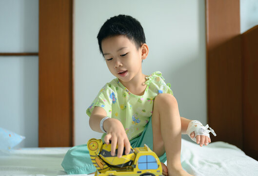 Young asian boy in hospital gown playing toy car while sitting on hospital bed with IV catheter on his hand, positive representation of children in medical care. recovery and child patient concept - Powered by Adobe
