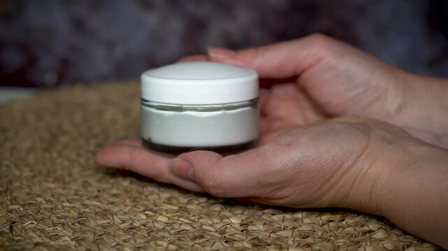 Woman&rsquo;s hand holding a glass jar with homemade natural face or body cream. Minimalist skincare concept with organic cosmetic on beige and dark background, eco-friendly and clean beauty style.