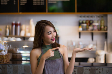 Young Asian female barista in apron smiling and pointing to her open palm, standing behind the coffee counter in cozy cafe, ready to serve customers or introduce products.