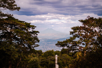 Panoramic view of a distant volcano and cityscape framed by lush tropical forest under a dramatic cloudy sky.

