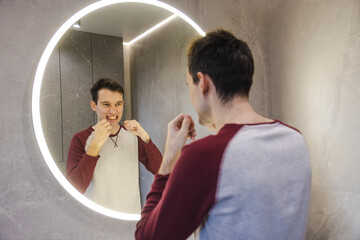 Young man flosses his teeth in front of a circular mirror, carefully maintaining oral hygiene as...