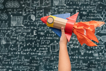 Hand Holding Toy Rocket with Flames in Front of Blackboard Filled with Math and Science Formulas