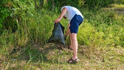 Man cleaning litter in tall grass with black garbage bag. Summer environmental activity in natural...