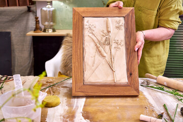 Person holding completed botanical plaster relief in wooden frame. Handmade floral artwork with...