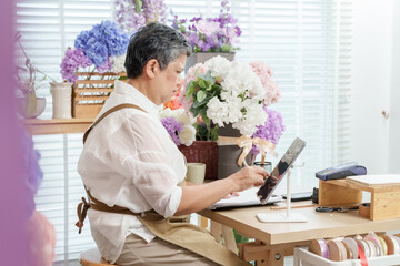 Asian senior female florist using tablet while sitting at wooden desk surrounded by flower arrangements in shop working on online orders digital platform modern small business after retirement