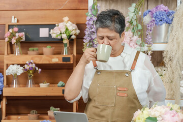 Asian senior florist closing eyes while drinking warm tea in peaceful workspace with flower bouquet...