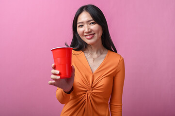 Asian woman with long black hair in orange dress smiling while extending a red plastic cup toward...