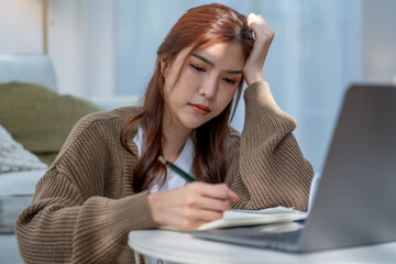 woman with long hair is sitting at table, looking stressed while writing in notebook next to...