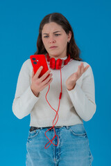 Concerned young woman in white sweater holding red smartphone with headphones, reacting in shock...