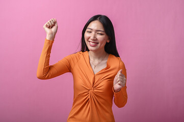 Asian woman with long black hair in orange dress smiling and dancing joyfully with raised arms, standing indoors against pink background, expressing fun, energy, and cheerful positive emotion