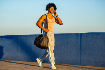 Stylish mixed race man walking along seaside promenade while talking on phone and carrying gym bag