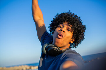 Naklejka premium Excited mixed race young man taking selfie at the beach with raised arm and headphones around neck