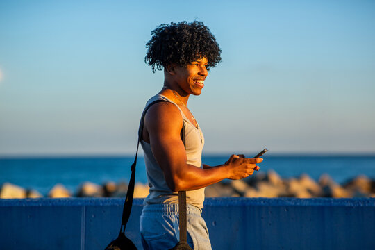 Smiling young man with athletic build using smartphone near ocean at sunset