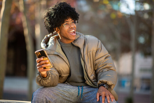 Young man with black curly hair laughing while holding smartphone outdoors in winter clothes