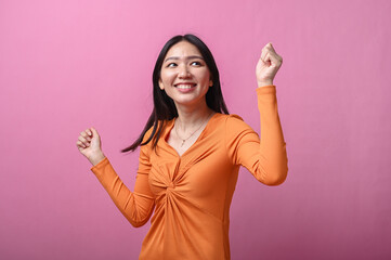Asian woman with long black hair in orange dress smiling and dancing joyfully with raised arms,...