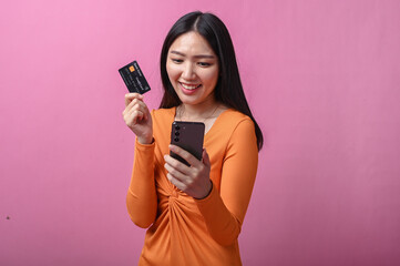 Naklejka premium Asian woman with long black hair wearing orange dress holding smartphone and credit card, standing indoors against pink background, smiling slightly while making an online payment or transaction
