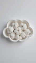 A cloud shaped bowl filled with fluffy cotton balls on a plain white surface top down view studio shot
